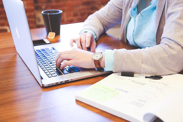 A headless individual types at a computer with a coffee and open book nearby